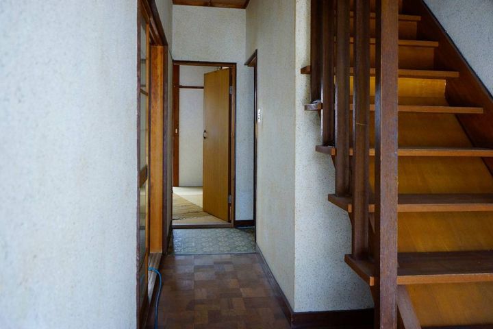 古い木造住宅の玄関。右手に階段があり、奥へ続く廊下と開いた室内ドアが見える。