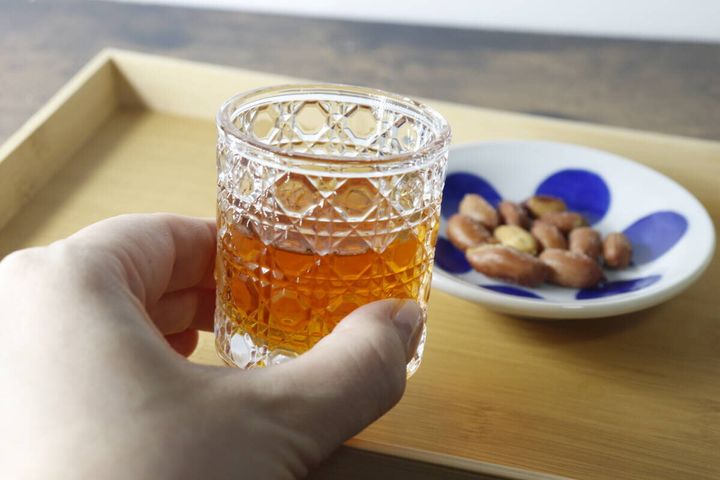 ミニグラスに入った琥珀色の飲み物と、おつまみのピーナッツを持つ手