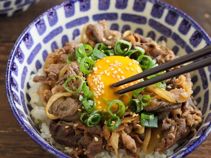 模様の丼のスタミナ丼に、黒い箸で中央の卵黄を崩そうとしている様子