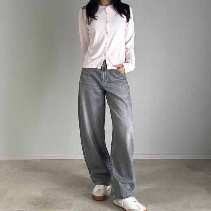 ピンクのカーディガンとグレーデニムを着用した女性が立つ
