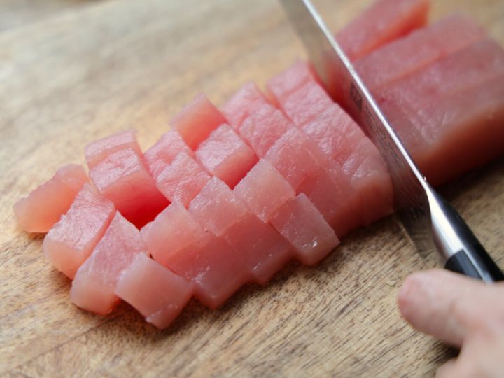 木のまな板の上で、包丁を使って刺身用のまぐろを2cm角に切っている手元。