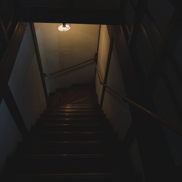 薄暗い古い木造住宅の階段。天井の照明が一つだけ灯り、奥へ階段が続いている。