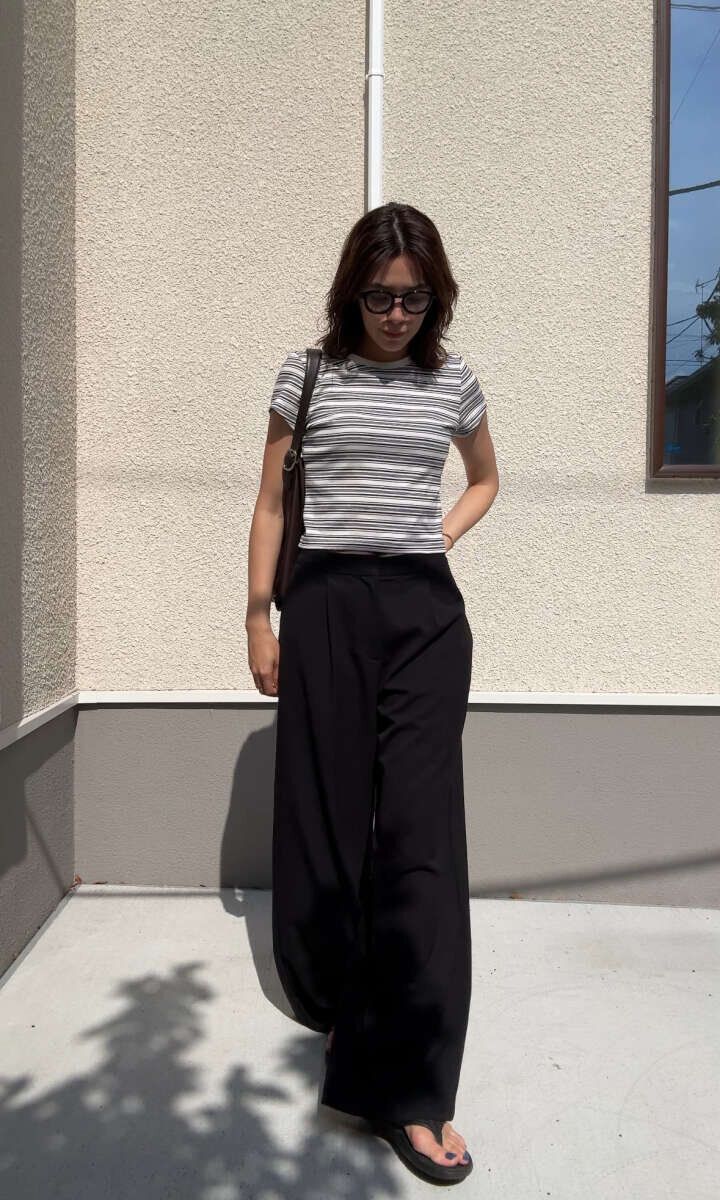 ボーダー柄のミニTと黒いワイドパンツを着用し、サングラスをかけた女性が立っている