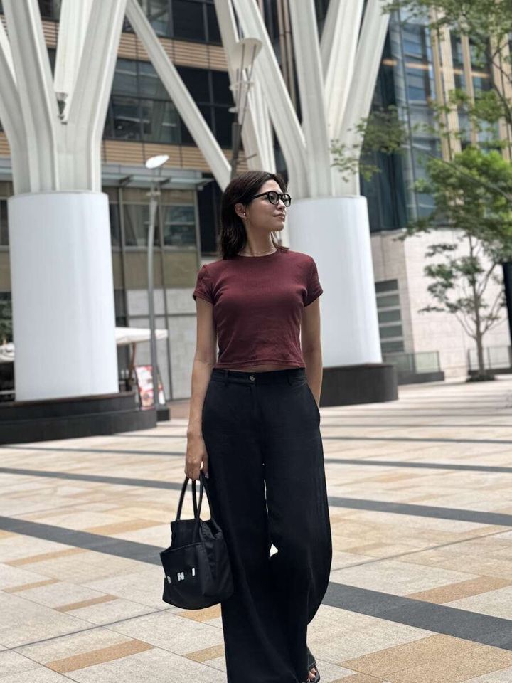 赤茶色のミニTと黒いワイドパンツを着用した女性が、街中でバッグを持って立っている
