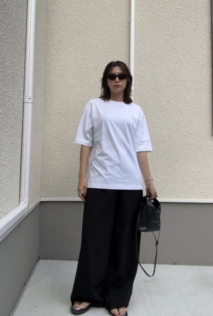 白いTシャツと黒いワイドパンツを着用し、サングラスをかけた女性がバッグを持って立っている