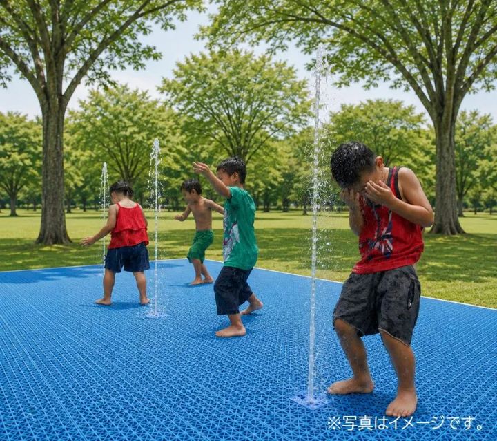 芝生と並木に囲まれた屋外で、子どもたちが青い床面から上がる細い噴水で水遊びをしているイメージ
