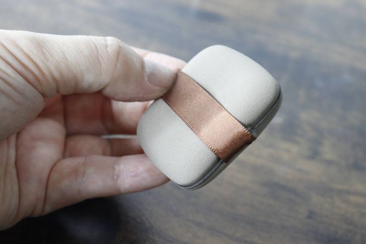 女性の手が、茶色いリボンが付いた四角いメイクパフを掴んでいる様子