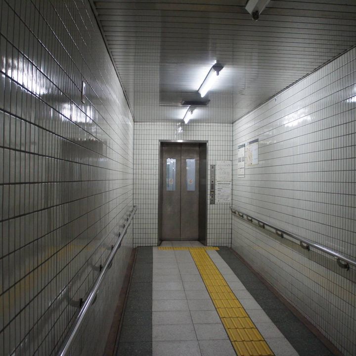 白いタイル張りの地下通路の奥にあるエレベーター。床の黄色い点字ブロックが扉の前まで続いている。