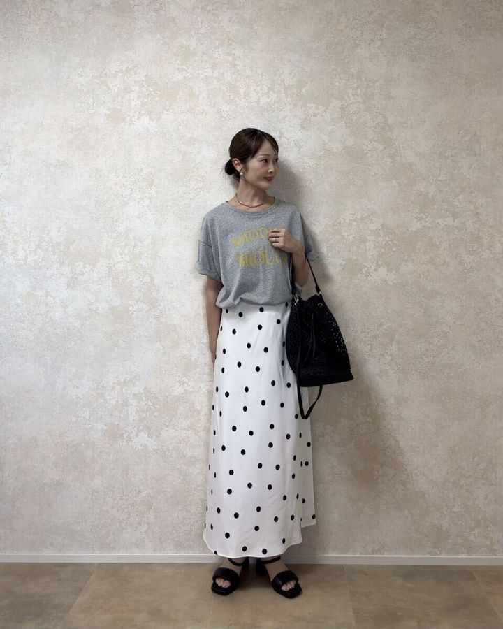 白のドット柄スカートとグレーのロゴTシャツを着た女性が斜めを向いて立っている