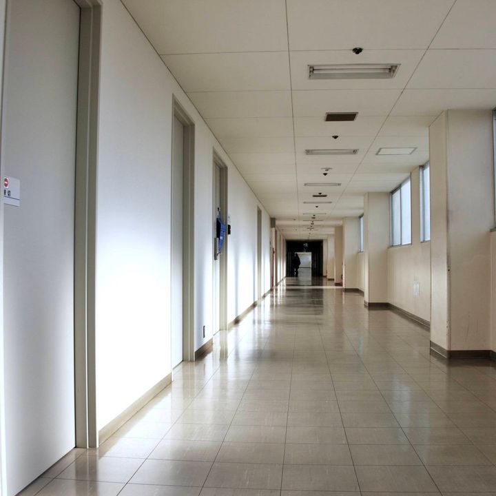 白い壁と窓が続く建物の長い廊下。廊下の奥には小さな人影が見え、突き当たりは薄暗くなっている。