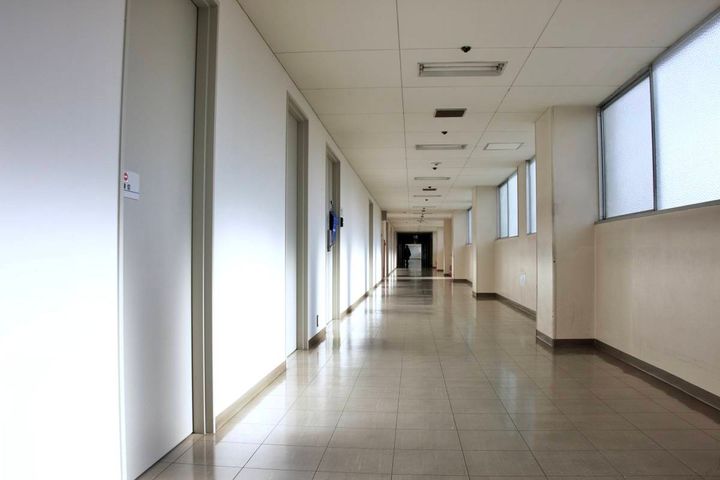 白い壁と窓が続く建物の長い廊下。廊下の奥には小さな人影が見え、突き当たりは薄暗くなっている。