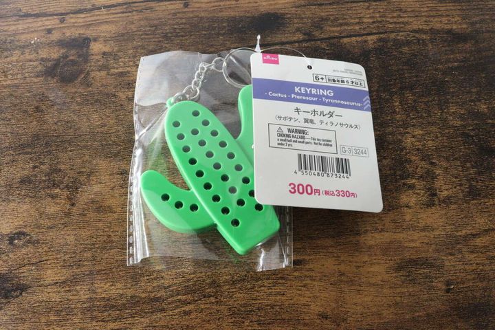 透明な袋に入った緑色のサボテン型キーホルダーと商品タグが木目のテーブルに置かれている