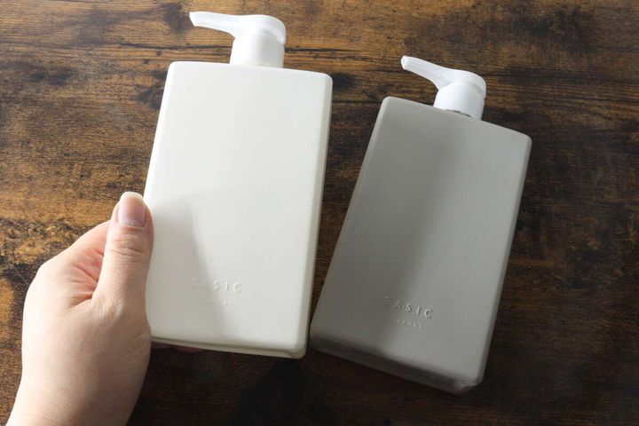 女性の手が白いポンプボトルを持ち、グレーのボトルと共に木目のテーブルに置かれている
