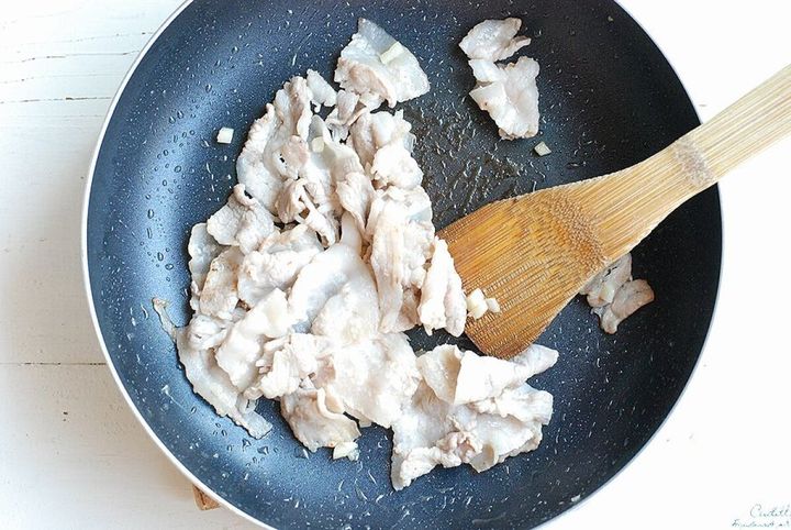 フライパンで白っぽくなるまで炒められている豚バラ肉と木製のへら