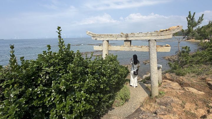 海に面して立つ石造りの鳥居とその下に立つ女性