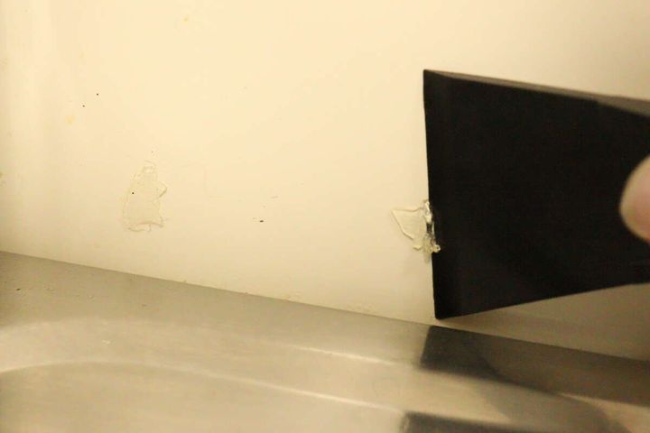 白い壁に付着した粘着汚れを40mm幅のヘラで剥がしている作業中の様子