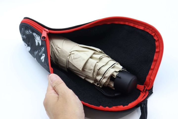 手が折りたたみ傘用収納袋にベージュの折りたたみ傘を入れようとしている。内側の黒い生地が見える。
