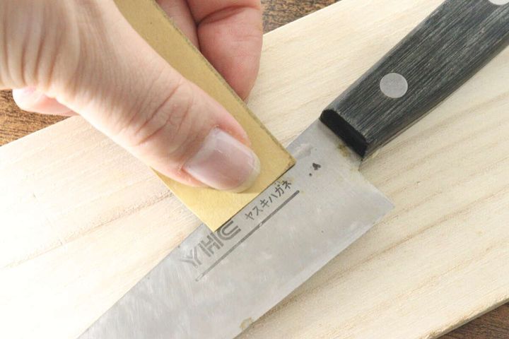 木製のまな板の上で、サンドペーパーを使って包丁のサビを研磨している手元