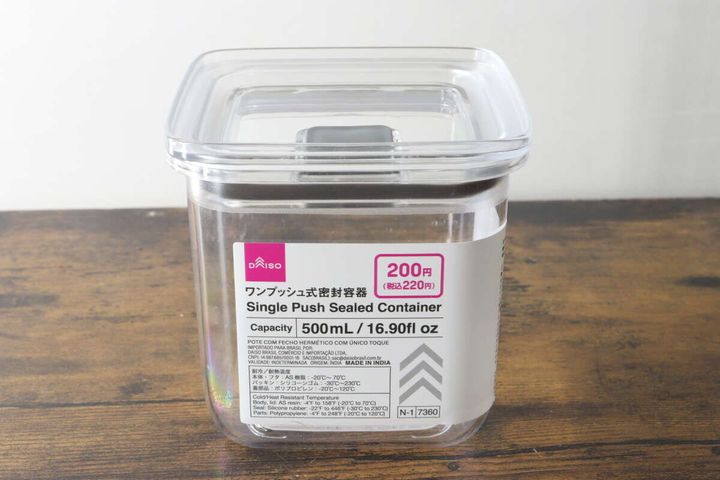 透明なワンプッシュ式密閉容器が木目のテーブルに置かれ、商品情報ラベルが貼られている