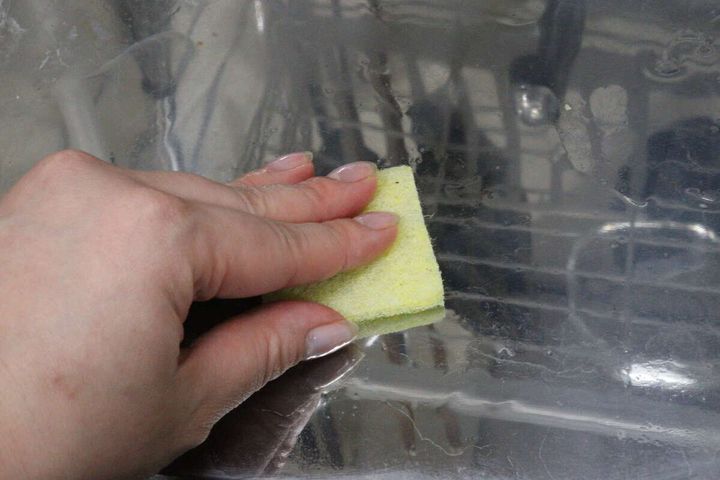 女性の手が黄色いクリーナーで水垢のついた水切りラックのトレーを擦っている