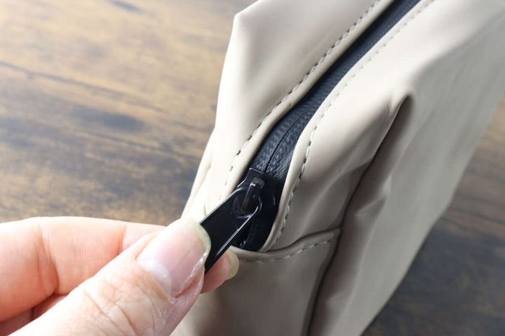 指でベージュのポーチの黒いファスナーを開けようとしている