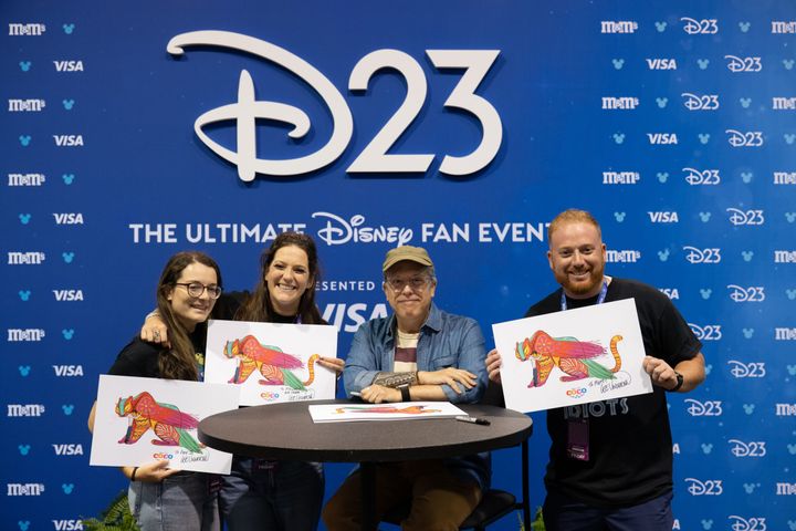 D23のバックドロップの前で、サイン入りのペピータのアートワークを掲げる4人のファンとリー・アンクリッチ