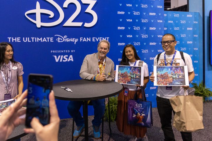 D23のバックドロップの前で、サイン入りの印刷物を持つ3人のファンと並ぶエリック・ロビソン