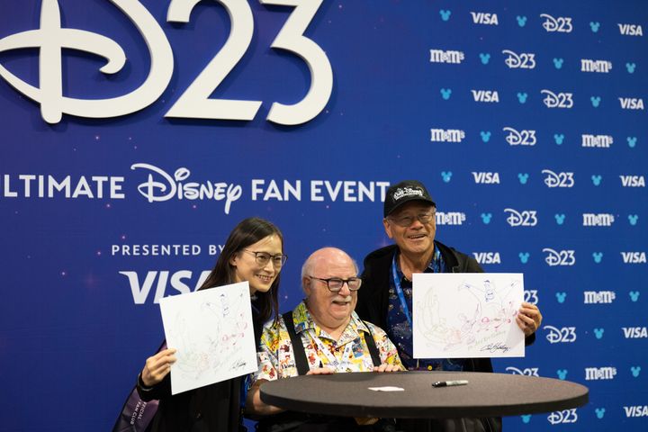 D23のバックドロップの前で、サイン入りのイラストを掲げた2人のファンと並ぶエリック・ゴールドバーグ