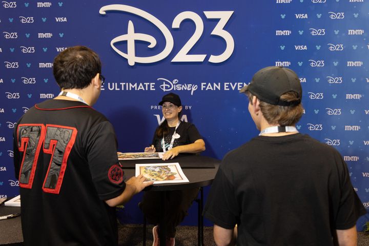 大きなD23のロゴを背にしたテーブルで、来場者のサインに応じるシェリー・マーティン