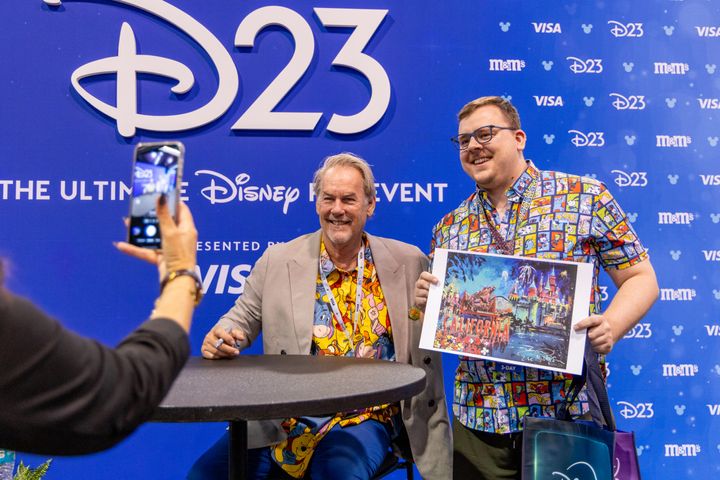 D23の大型ロゴを背に、「CALIFORNIA」と題したイラストにサインを入れるエリック・ロビソンと、印刷物を掲げるファン