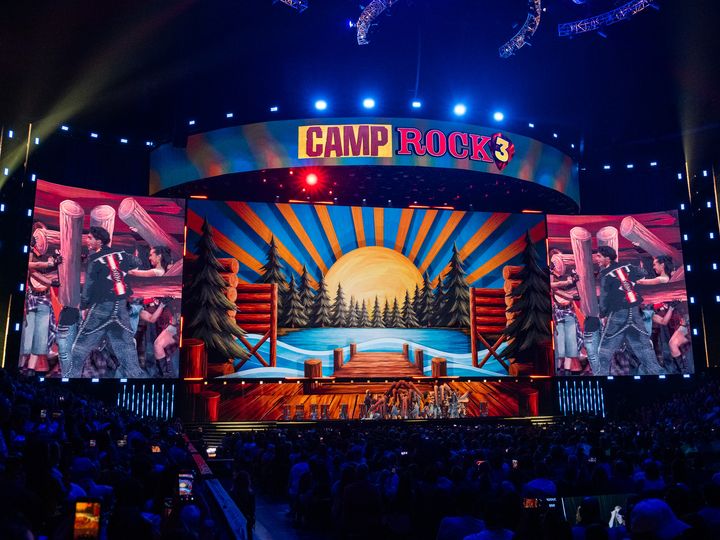 湖と桟橋を描いたキャンプのセットで歌う出演者たちとCAMP ROCK 3のロゴ