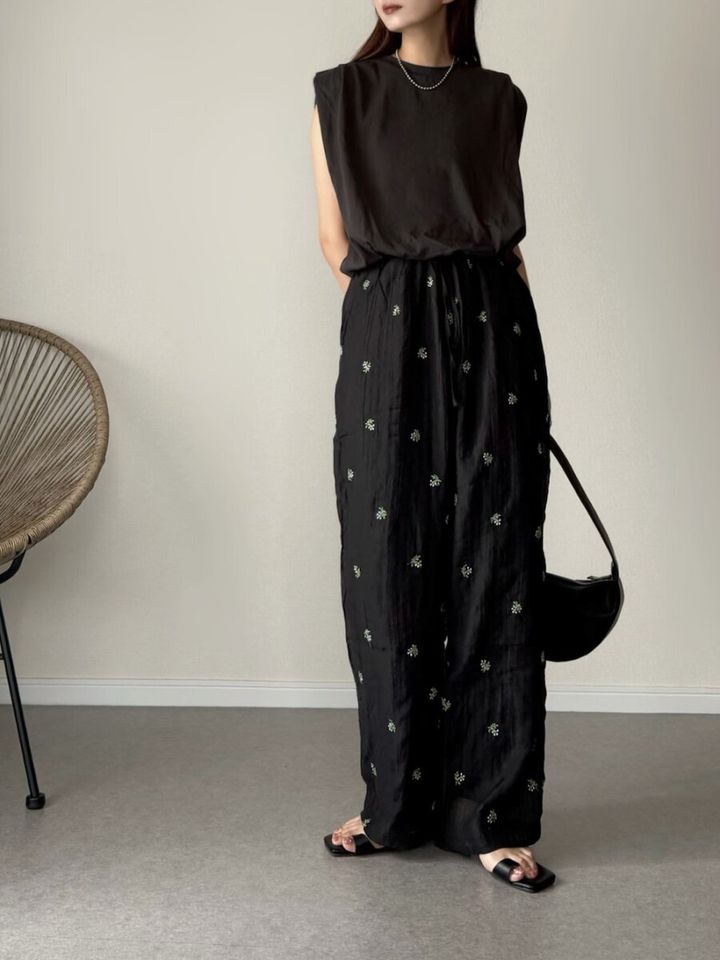 黒いノースリーブトップスと刺繍入り黒パンツを着た女性が立つ