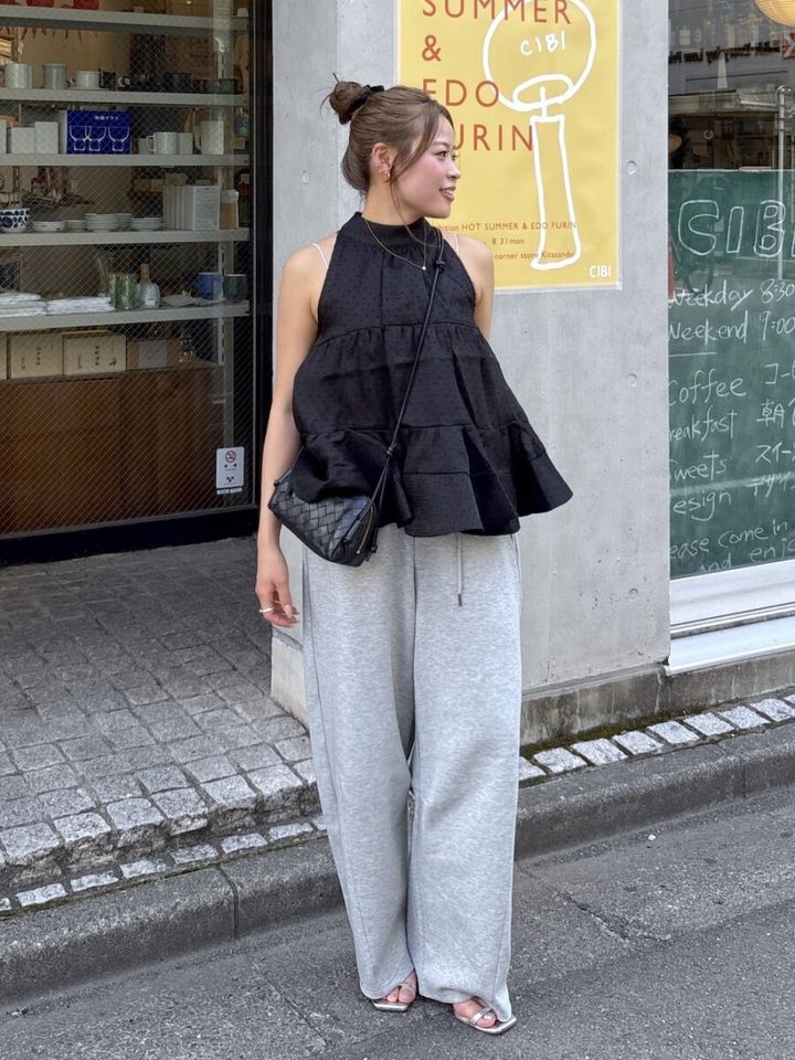 黒いティアードブラウスとグレーのスウェットパンツを着た女性が横向きに立つ
