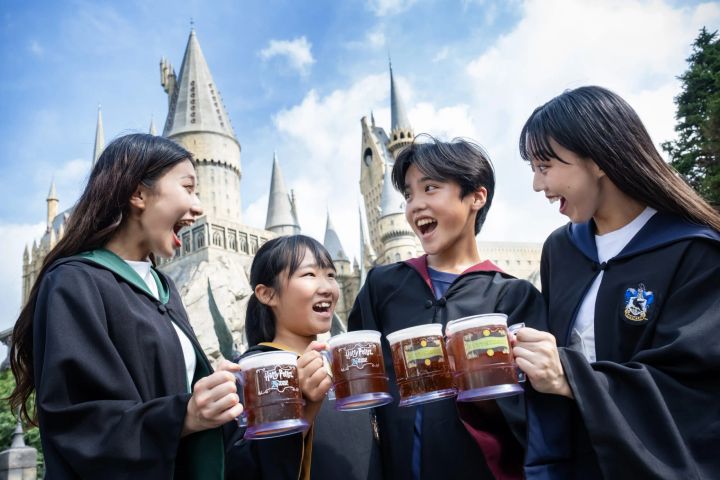 ホグワーツ城の前で25周年記念マグカップのバタービールを掲げるローブ姿の4人