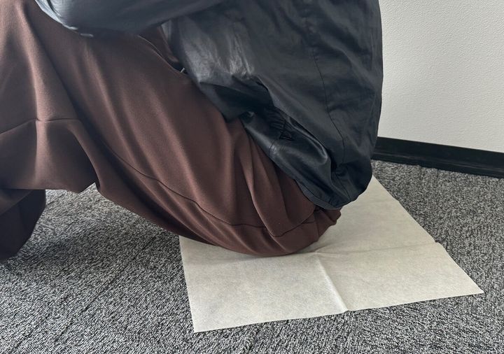 室内で黒い上着と茶色いパンツの人がレジャーシートに座っている様子