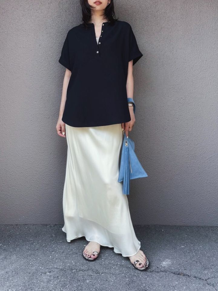 黒ブラウスと白サテンスカートの女性が青いバッグを持ち壁際に立つ