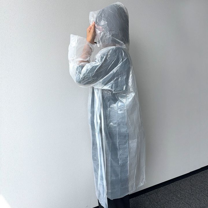 白い壁を背景に、透明なレインポンチョを着用した人物が横向きに立っている