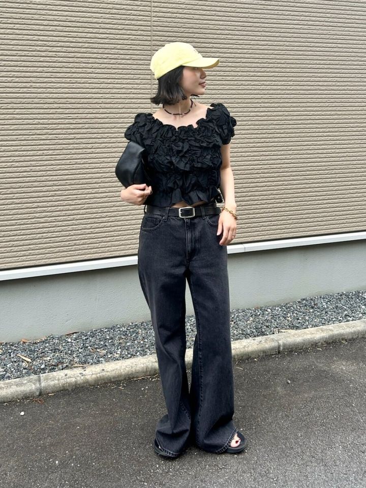 黒シャーリングブラウスと黒デニム、イエローキャップの女性が屋外に立つ