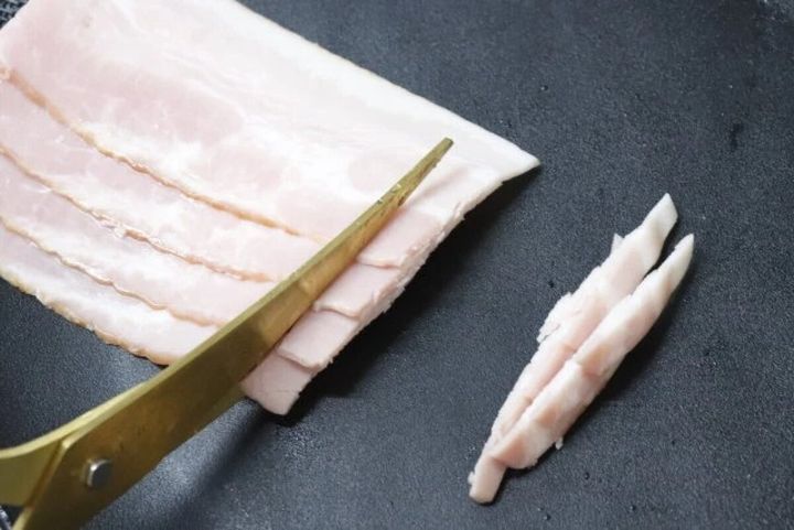 黒いフライパンの上で、金色のキッチンバサミが薄切りベーコンをカットしている