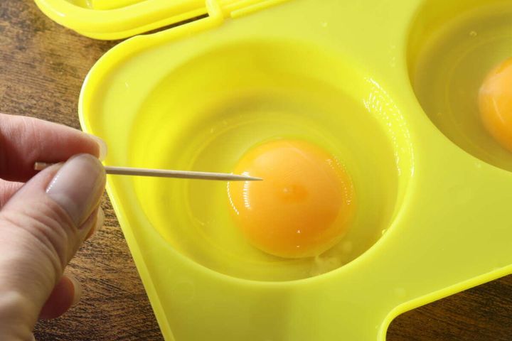 黄色い調理器のくぼみに割った卵が入っており、つまようじで黄身に穴を開けている手元。