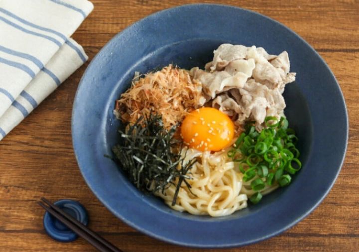 青い皿に盛り付けられたうどん、豚肉、卵黄、かつお節、刻み海苔の混ぜそば