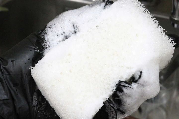 黒い手袋をはめた手で白い速乾キッチンスポンジを泡立てている。