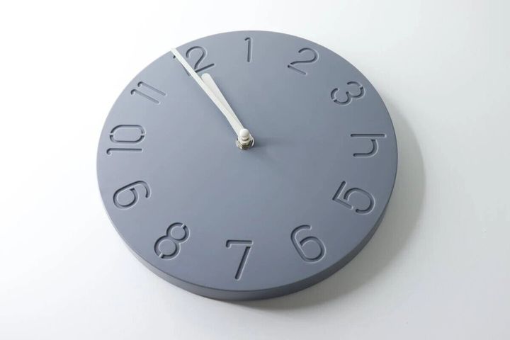 グレーのシンプルな掛け時計が白い背景に置かれ、白い針が12時を指している