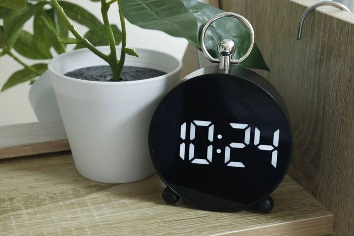 黒いLEDデジタル時計が木製の棚に置かれ、隣には観葉植物がある。時刻は10:24