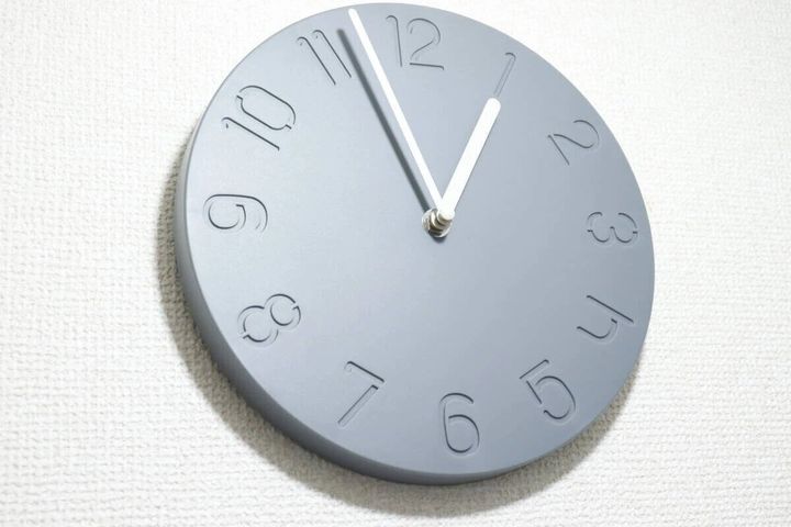 グレーのシンプルな掛け時計が白い壁に掛けられ、白い針が12時を指している
