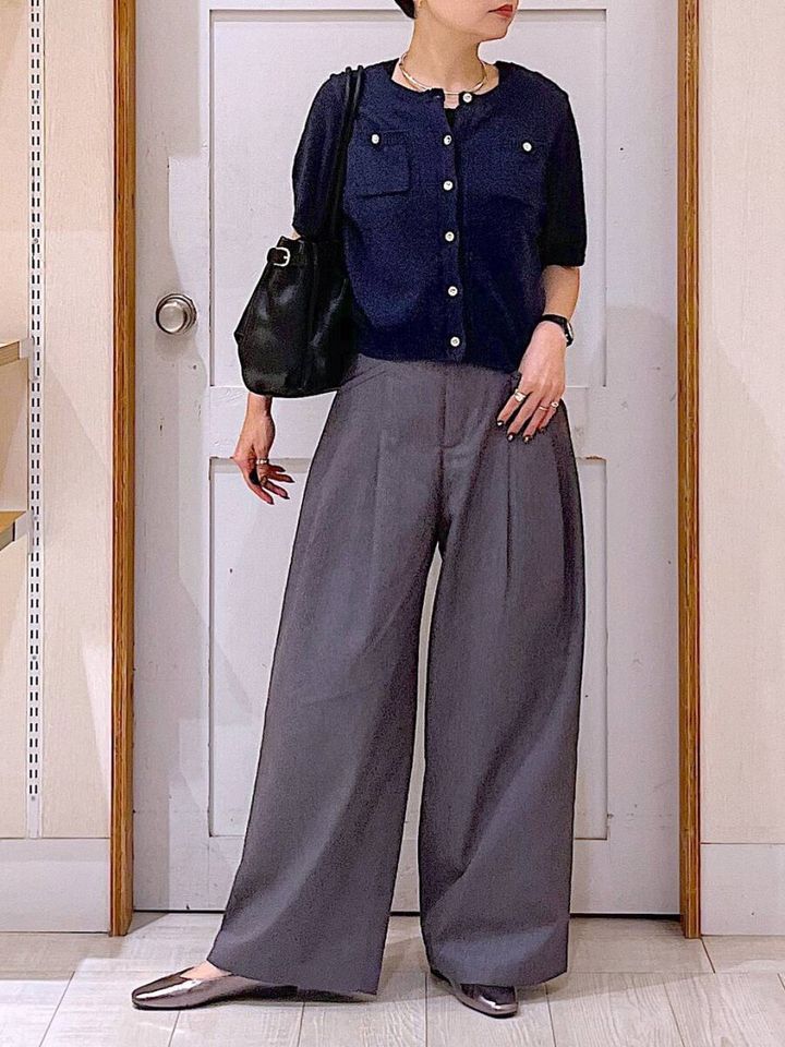 紺色の半袖カーディガンとグレーのワイドパンツを着用した女性が、黒いショルダーバッグを持ち、白いドアの前に立っている。
