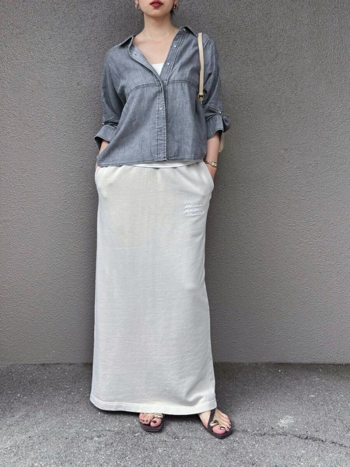 グレーのデニムシアーシャツと白のロングスカートを着用し、両手をポケットに入れている女性