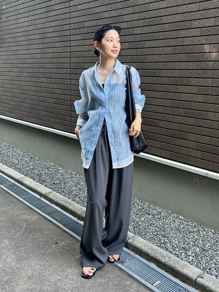 水色のストライプシアーシャツとグレーのワイドパンツを着用し、屋外で横を向く女性
