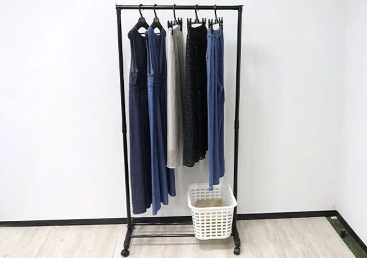 黒いハンガーラックに複数の洋服がかけられ、下に白いカゴが置かれている