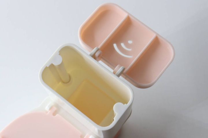 ピンクと白の便器型スマホスタンドのタンクのフタが開いており、中に小物収納スペースが見える